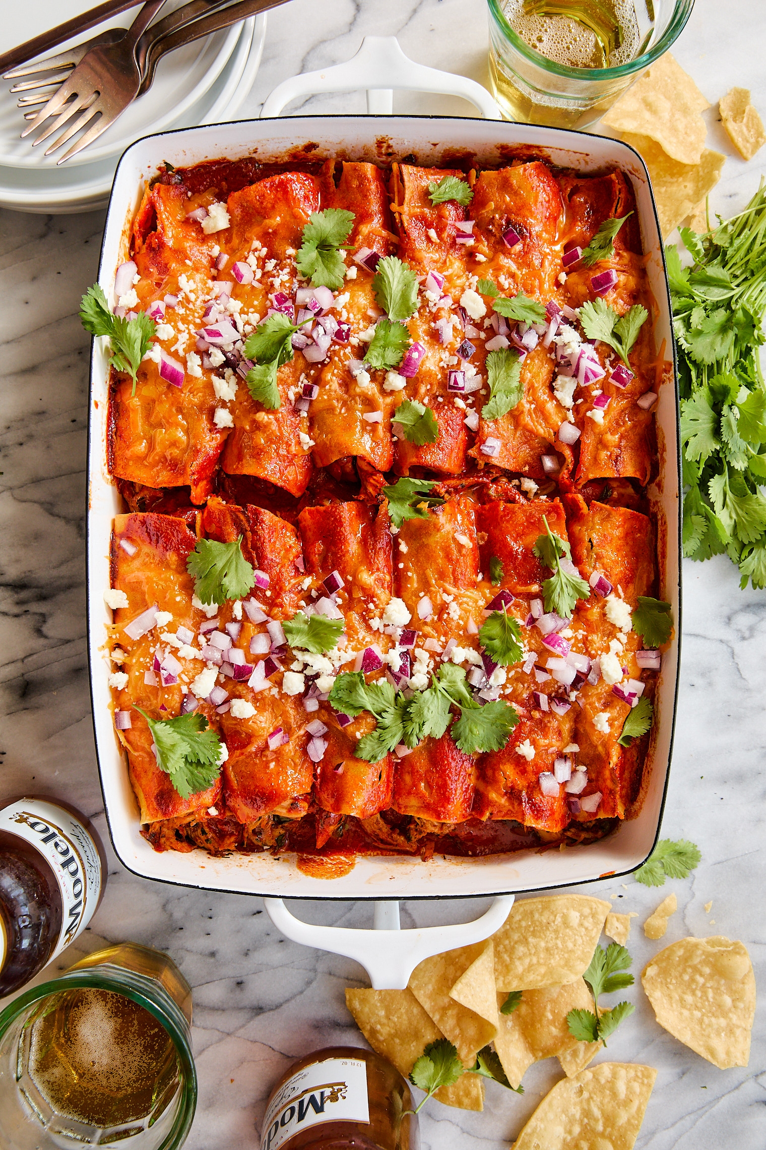 A casserole dish filled with enchiladas topped with cheese, red onions, and cilantro, surrounded by tortilla chips, beer bottles, and fresh cilantro