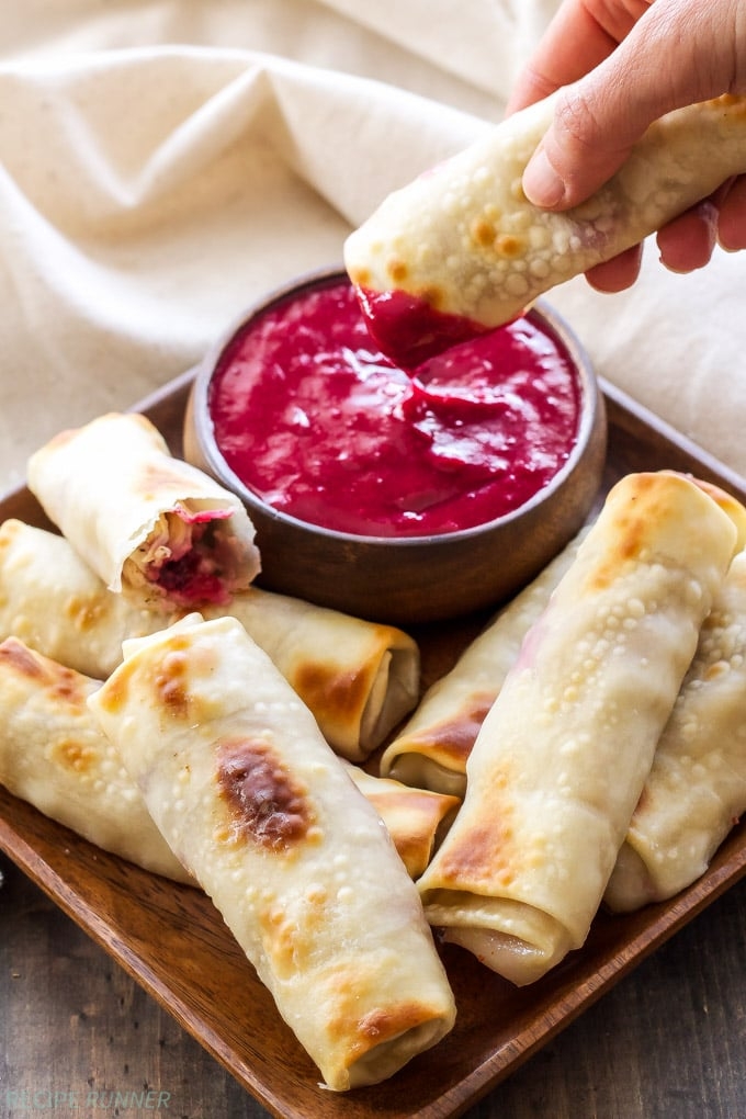 A hand dips a crispy egg roll into a bowl of bright red sauce, surrounded by more egg rolls on a wooden tray