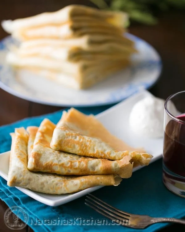 Thin crêpes folded on a plate with a glass of dark liquid and a dollop of cream in the background. More crepes are stacked on another plate