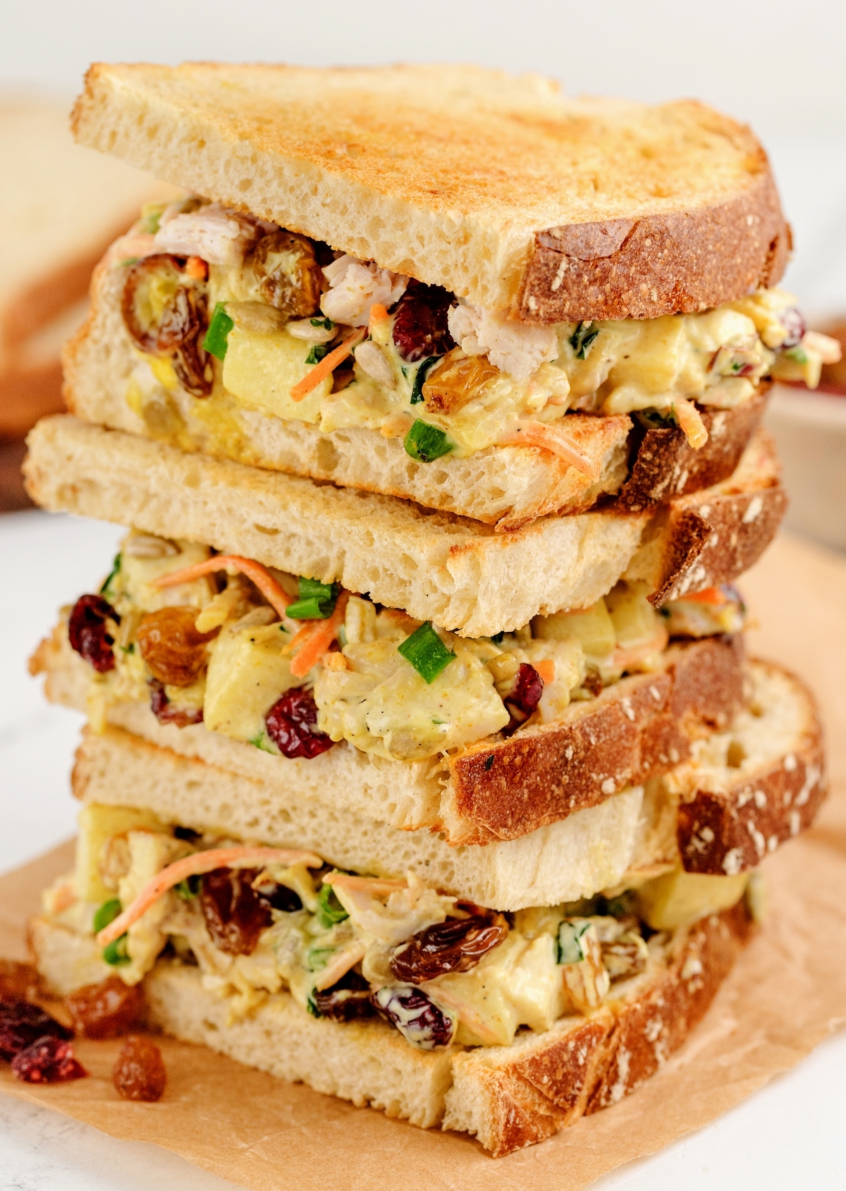 Stacked chicken salad sandwiches with grilled bread, featuring raisins, cranberries, and fresh herbs, placed on a wooden cutting board