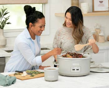 Two women successful a room utilizing a dilatory cooker, preparing a repast with caller ingredients, smiling and engaged successful cooking activities