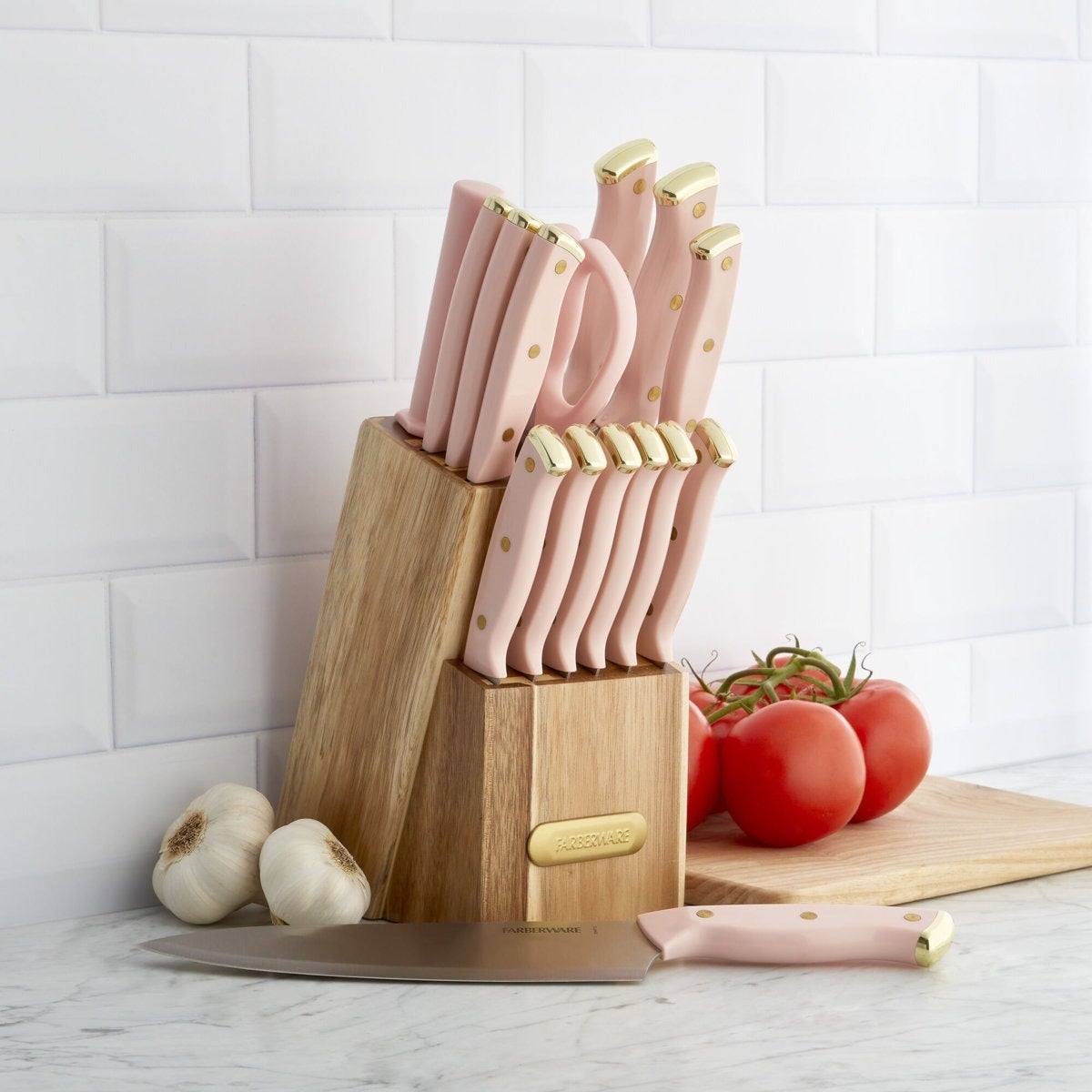 Knife set with pink handles in wooden block on kitchen counter, featuring tomatoes and garlic beside it
