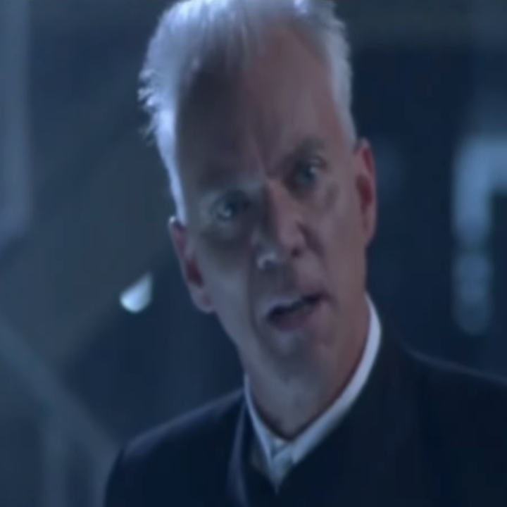An older person with white hair and a serious expression looks ahead, wearing a dark suit with a white collar