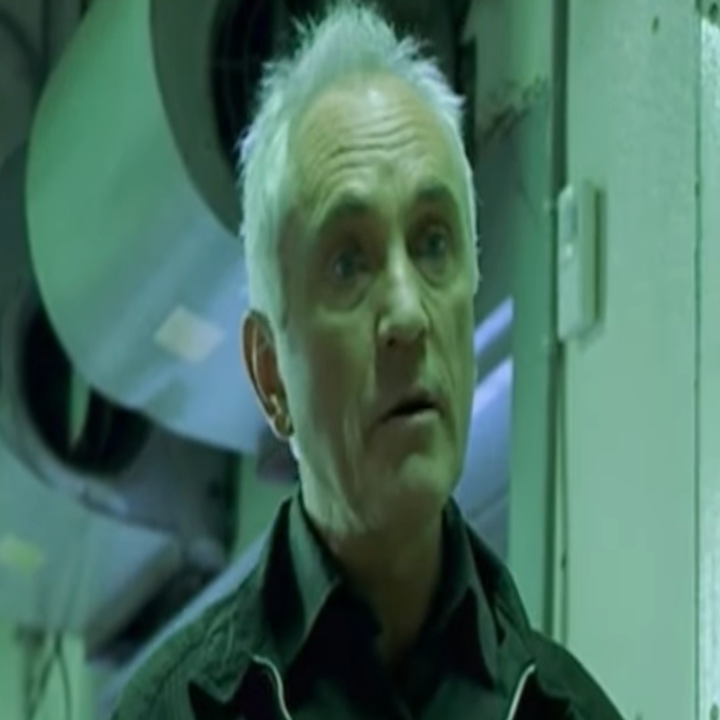 An older man with short white hair is speaking in an industrial setting with large pipes in the background