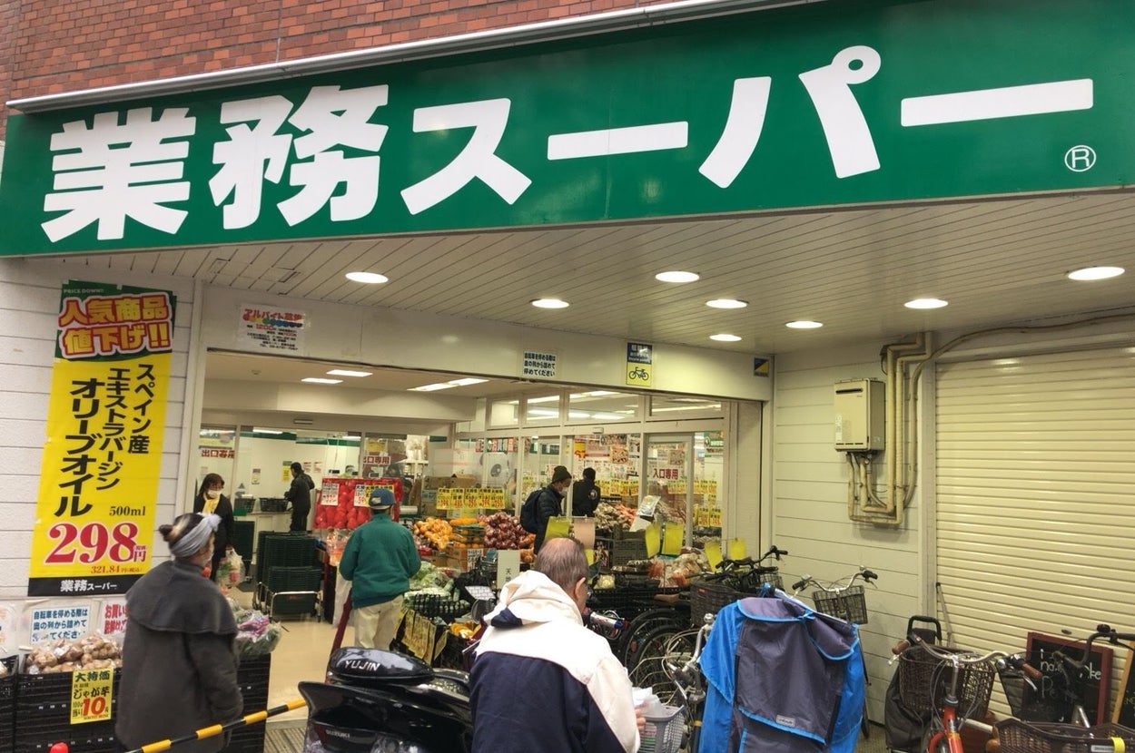 人々が出入りするスーパーマーケットの入り口。広告にはスペイン産オリーブオイルの特価情報が表示されています。