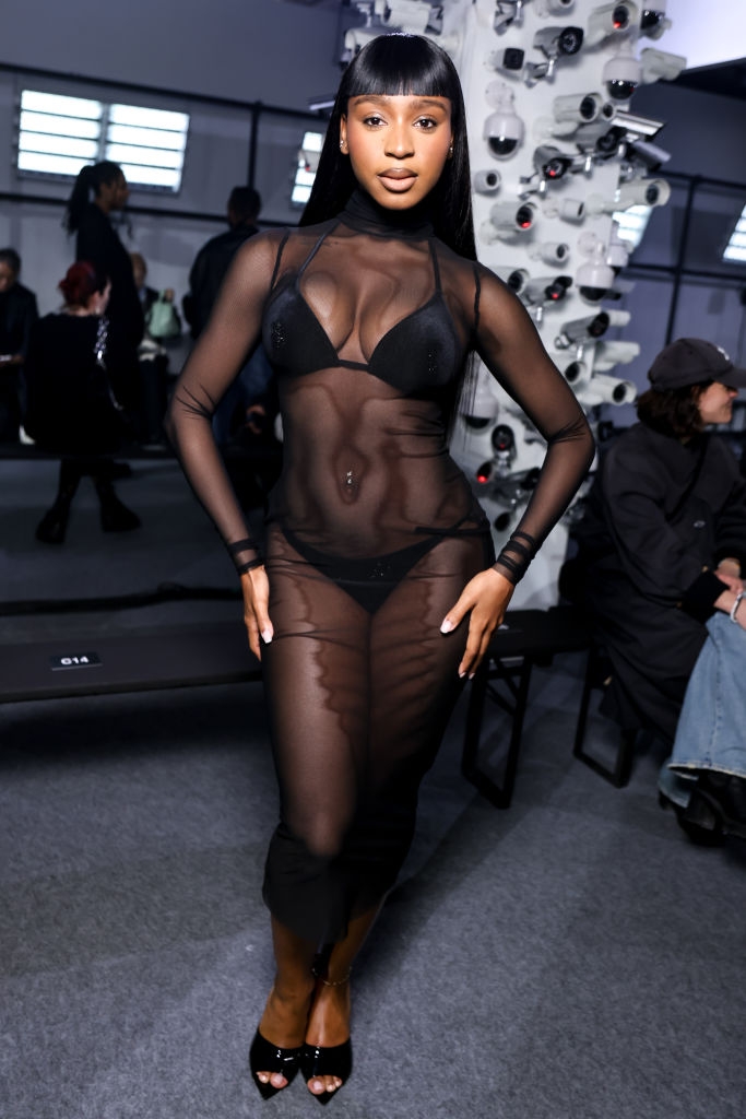 A person poses in a sheer black dress layered over a bikini at a fashion event, exuding style and confidence