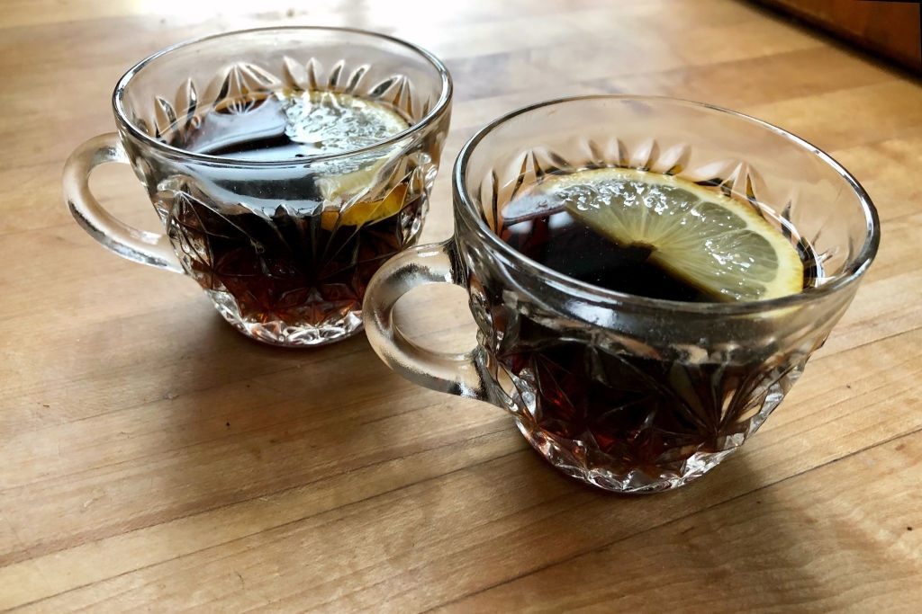 Two glass cups with a dark beverage, garnished with lemon slices, sitting on a wooden surface
