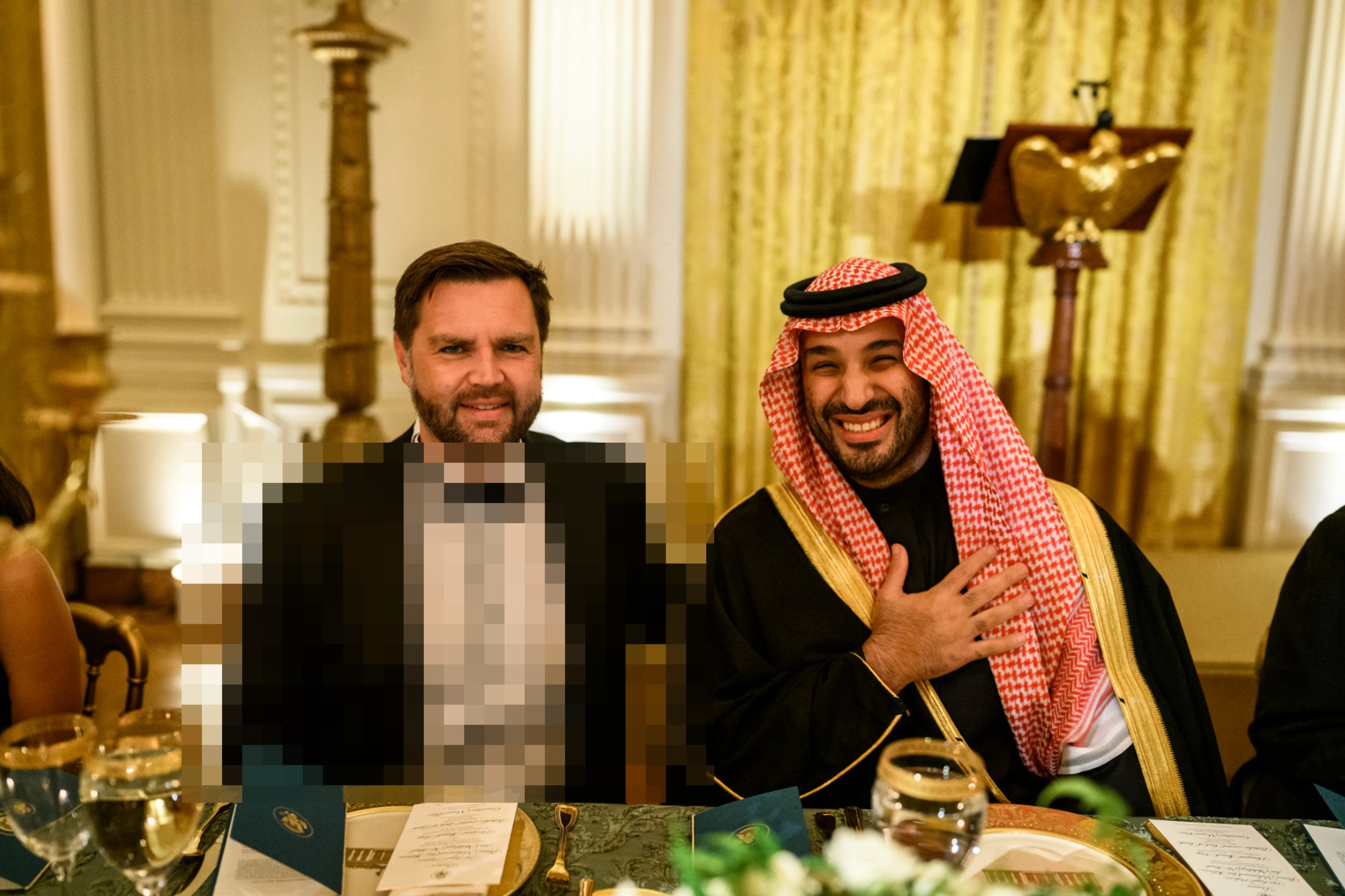 Two men sitting at a formal dinner table, one in a tuxedo and the other in traditional Middle Eastern attire, smiling and engaging warmly