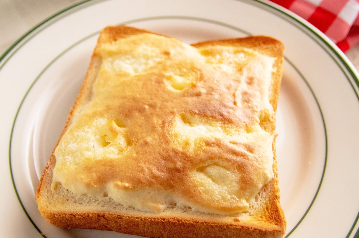 パンの上にふんわり焼かれたチーズトーストが皿に載っている。背景には赤と白のチェック柄の布がある。