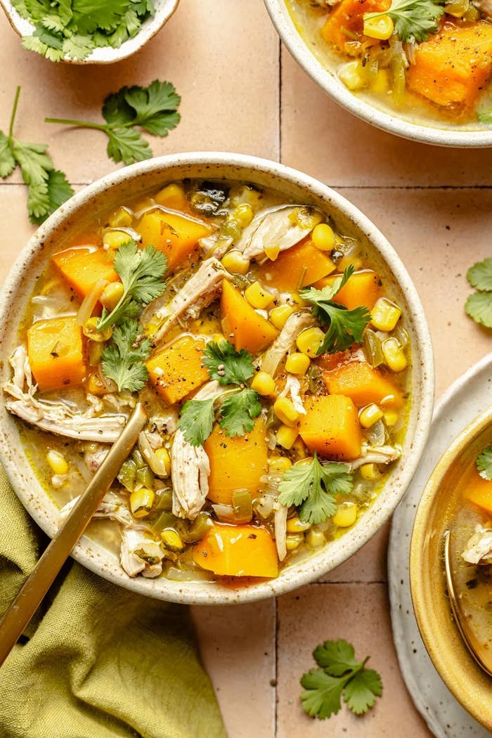 Bowl of chicken stew with corn, diced squash, and cilantro garnish, placed on a tiled surface. Nearby are a linen napkin and fresh cilantro leaves