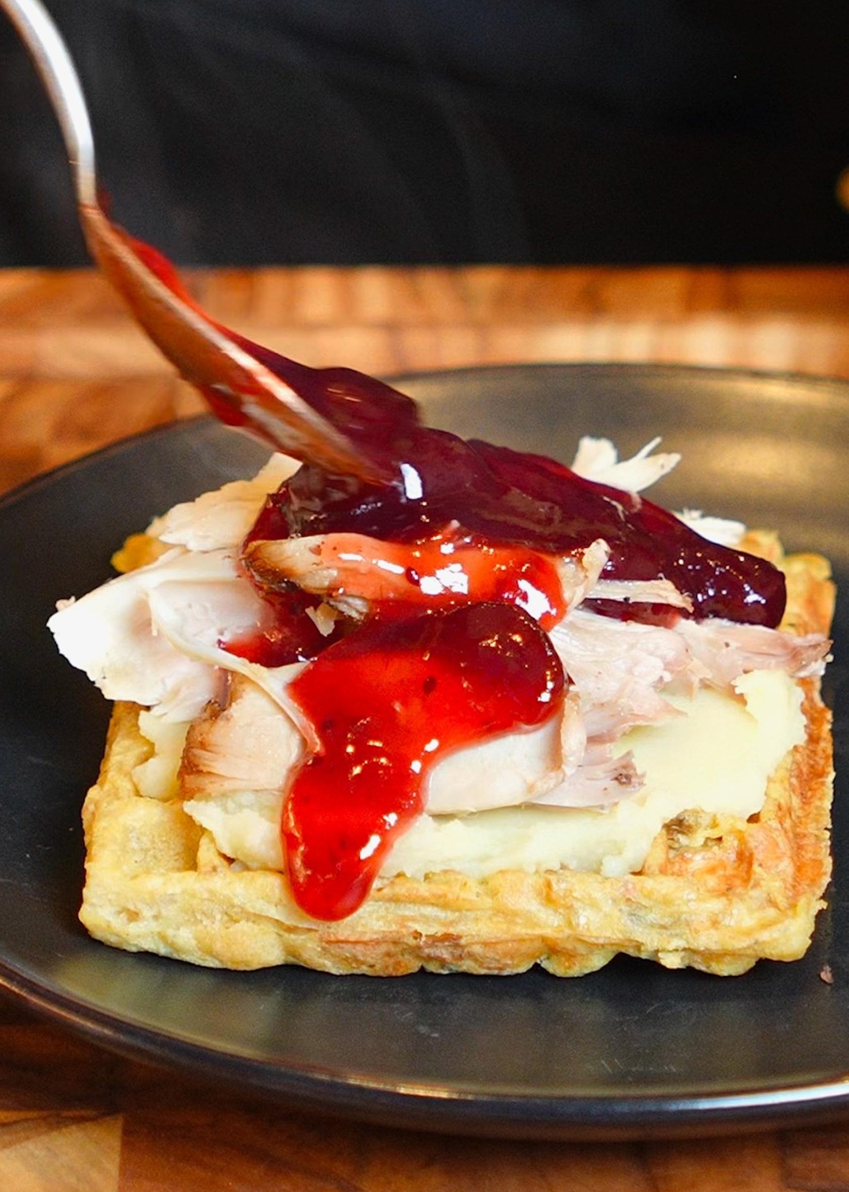 Waffle topped with turkey, cheese, and cranberry sauce. A spoon adds more sauce, on a black plate, viewed in a kitchen setting