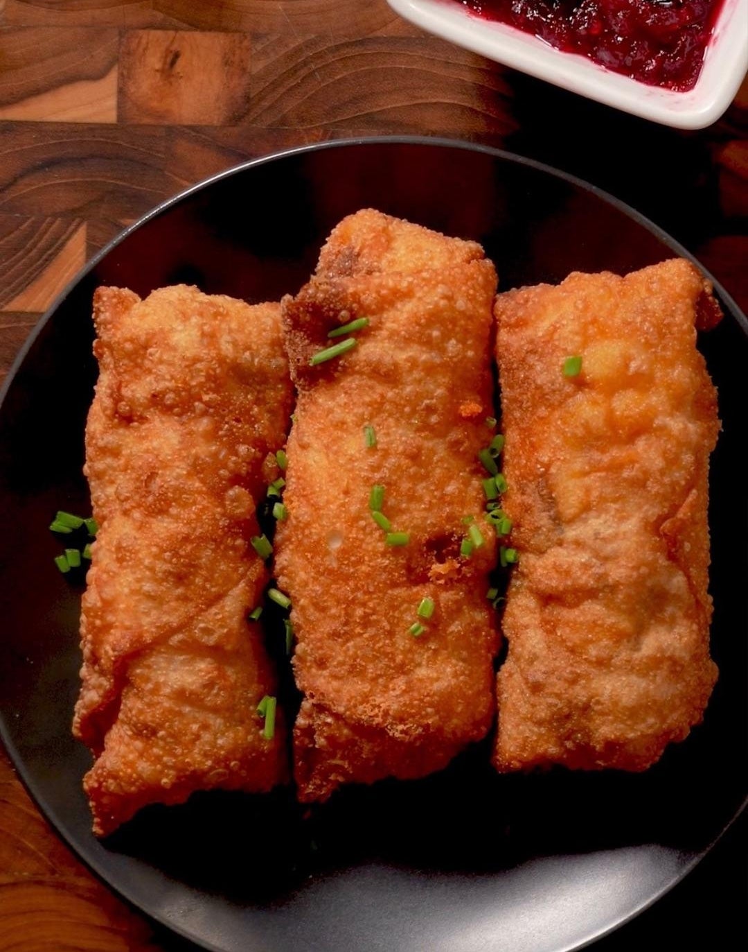 Three crispy egg rolls on a black plate, garnished with chives, served next to a dish of red berry sauce on a wooden table