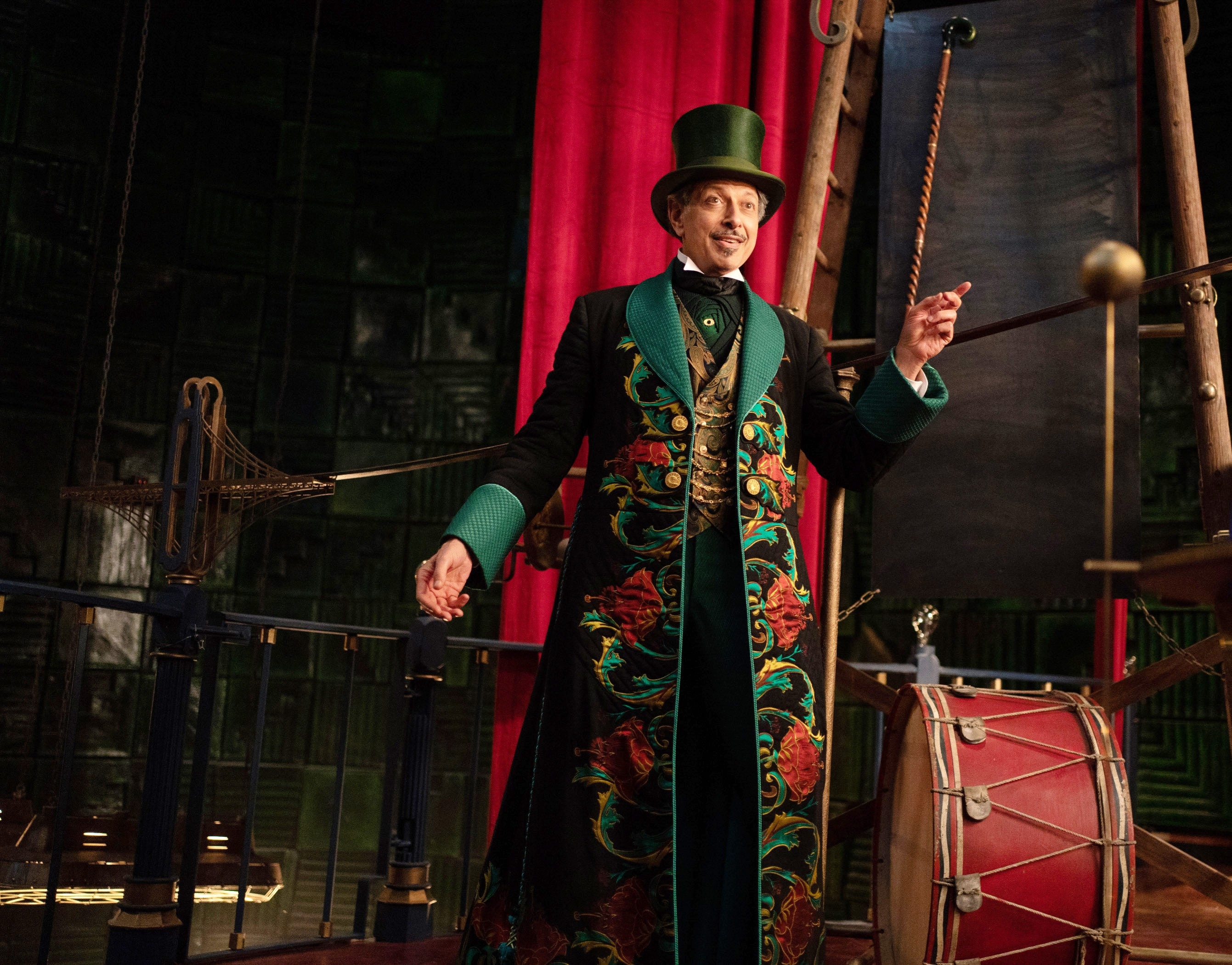 Person in a flamboyant Victorian-style coat and top hat on a theatrical set with a red curtain and drum, gesturing energetically
