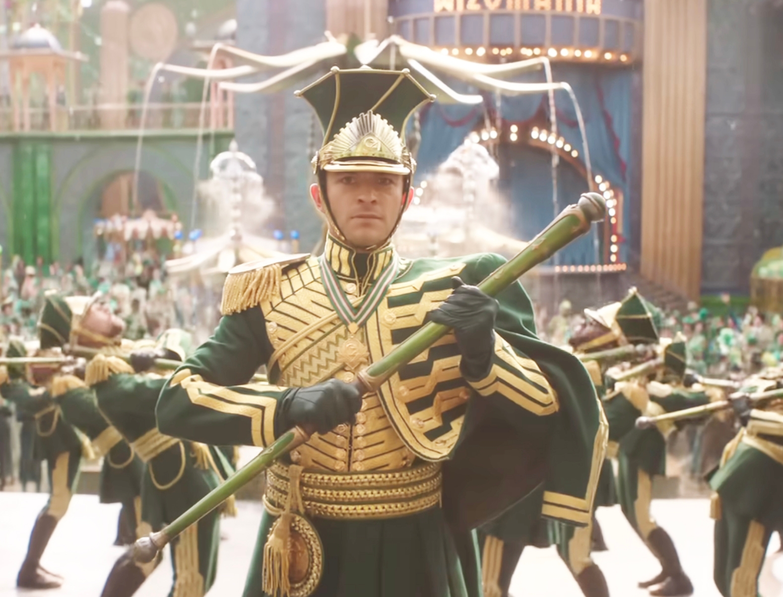 A performer in elaborate, military-style ceremonial attire holds a baton, leading a uniformed marching band in a theatrical setting