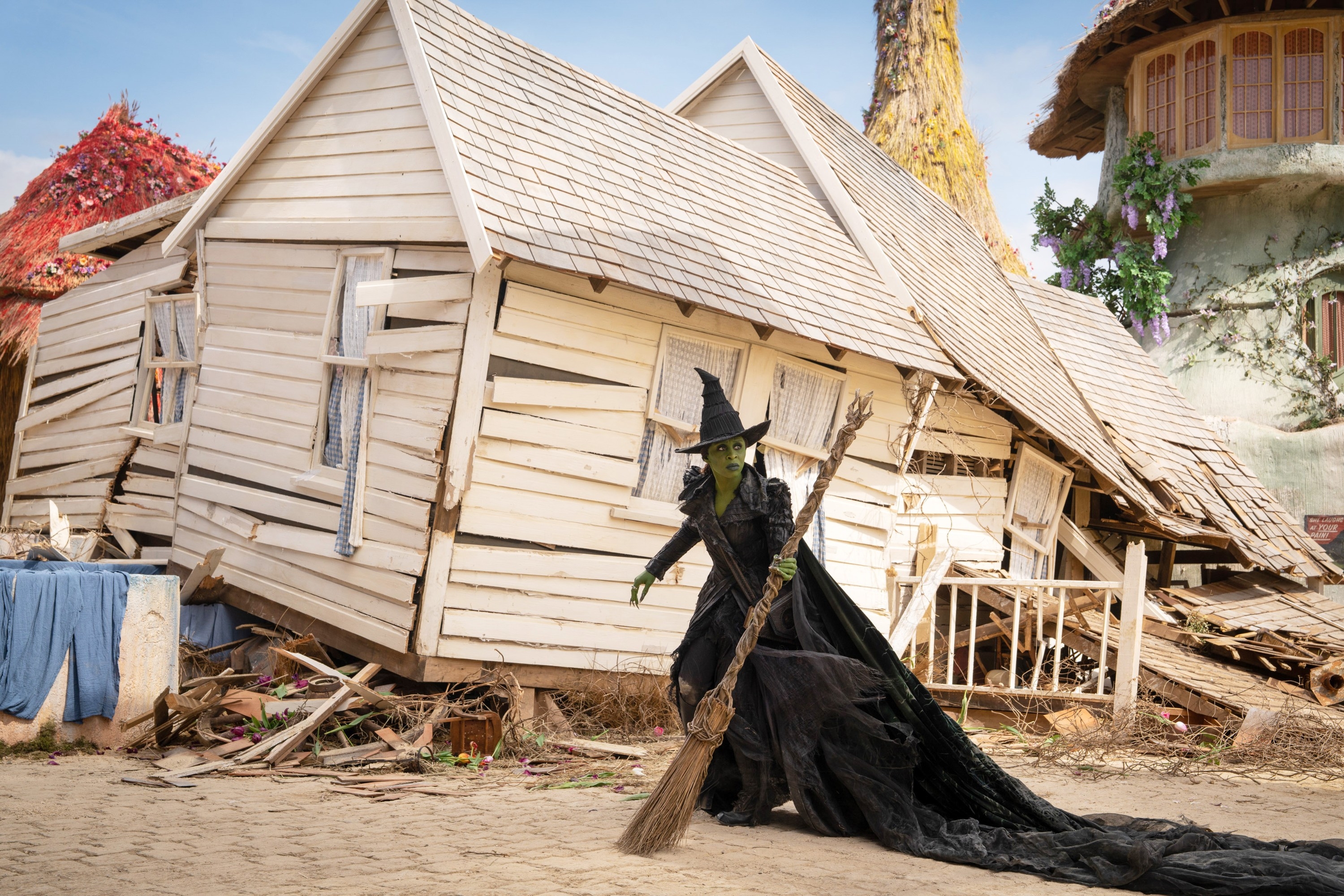 A witch in a black robe and hat stands with a broom in front of a tilted, ramshackle house