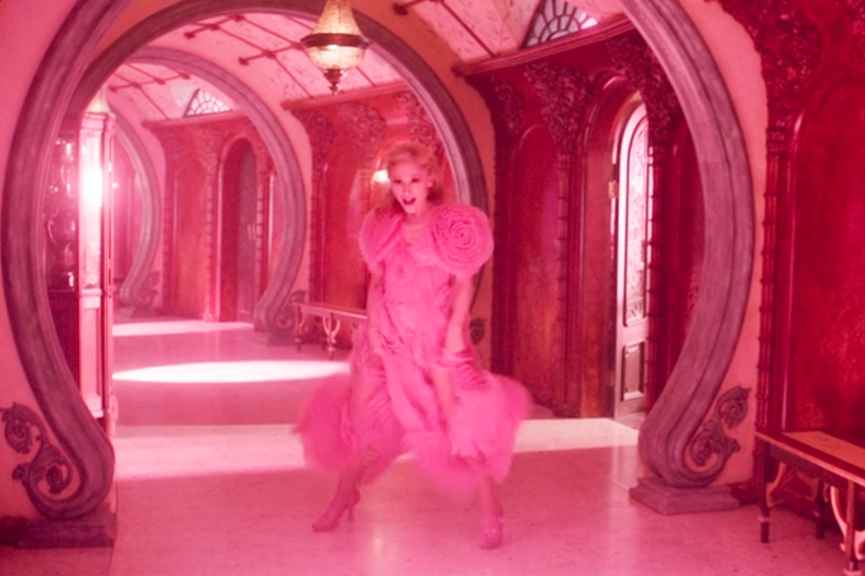 Person in an extravagant, flowing dress with large floral details, posed dramatically in an elegant arched hallway