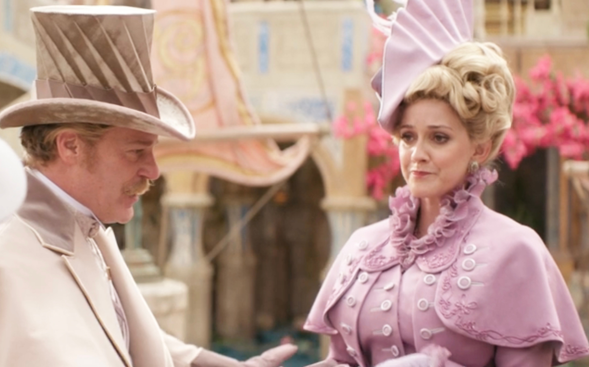 Two actors in period attire with elaborate hats in a historical or fantasy setting, appearing in a scene from a film or TV show