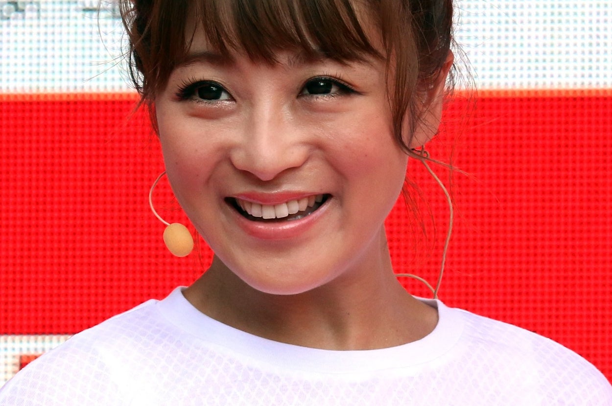 女性が白いトップスを着て笑顔で立っている。背景にオリンピック関連のロゴが見える。