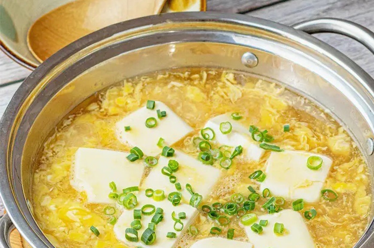 豆腐と卵が入った鍋料理。上に刻みネギが乗り、後ろには木のスプーンと器が見える。
