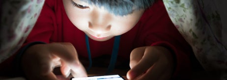 ベッドに隠れてスマートフォンを操作する子供。画面に集中し、指を動かしている。