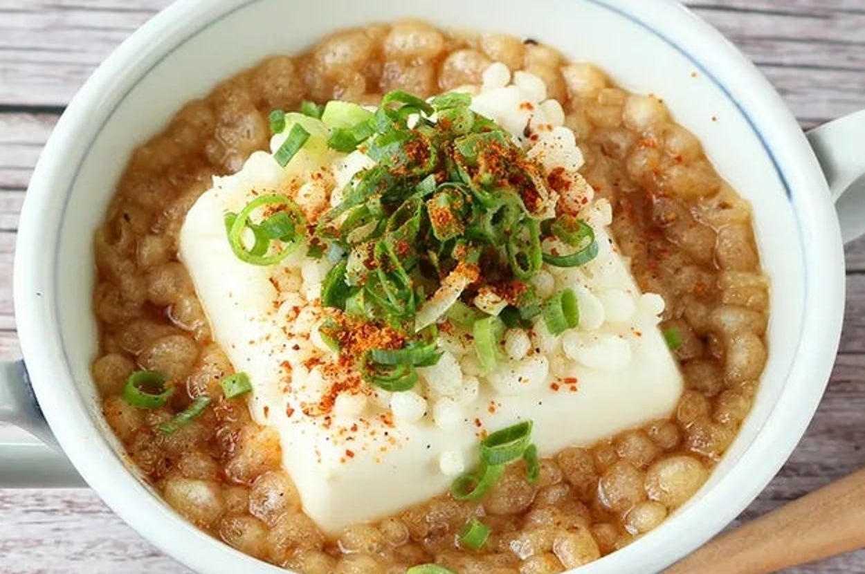 白い器に盛られた豆腐料理。豆腐の上にネギと七味が振りかけられ、揚げ玉がトッピングされている。横に木のスプーンがある。