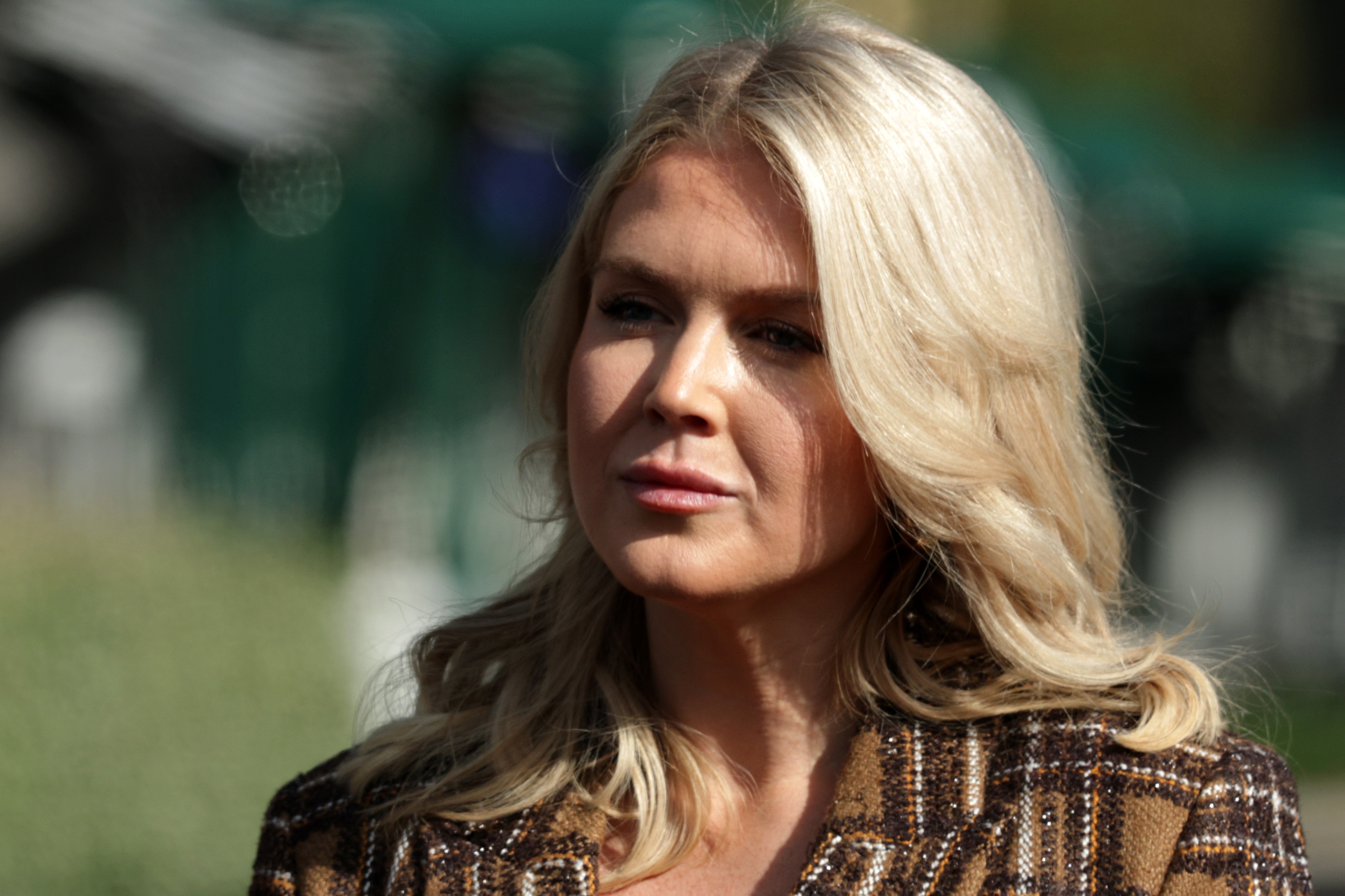 A person with long hair is standing outdoors, wearing a plaid coat