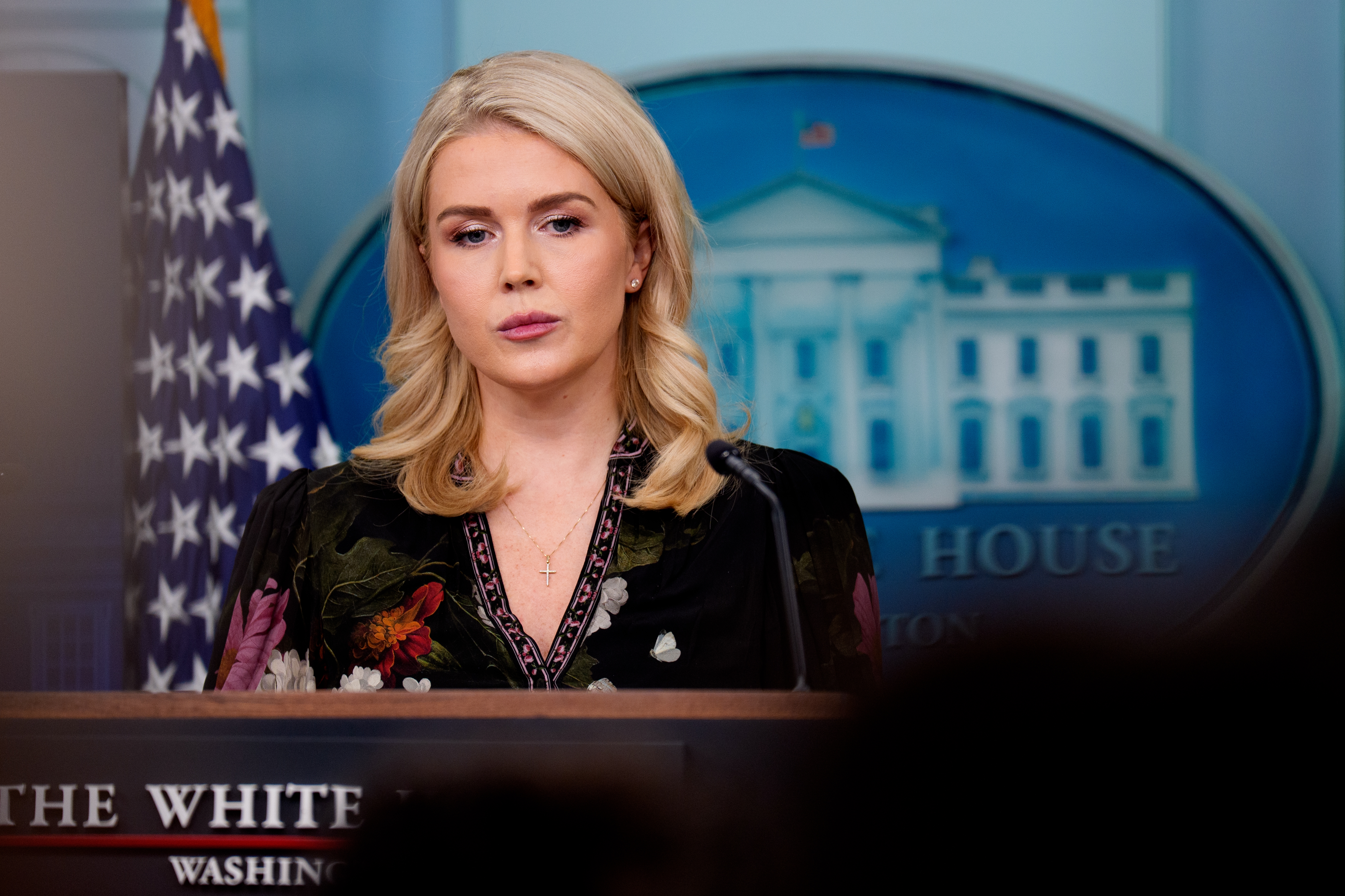 Person stands at a podium in a floral dress with a cross necklace, in front of a White House backdrop and U.S. flag