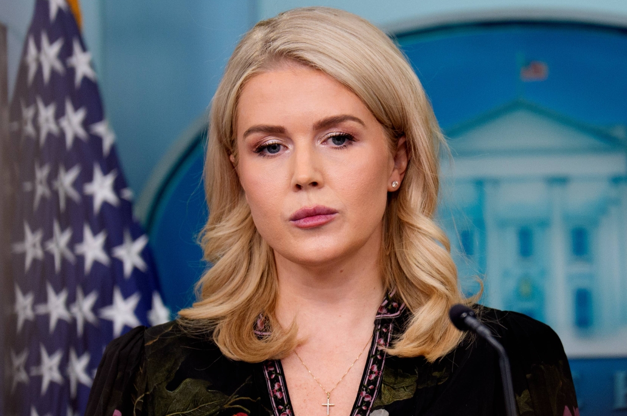 A woman speaks at a podium in a press briefing room, with a U.S. flag and a blurred government seal in the background