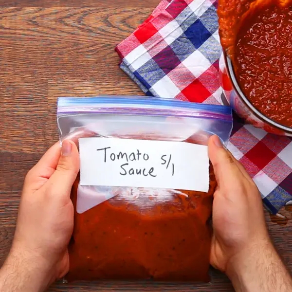 Hands holding a labeled freezer bag with tomato sauce on a wooden table, next to a pot of sauce and a checkered cloth