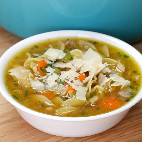 A bowl of noodle soup with veggies and shaved cheese on top