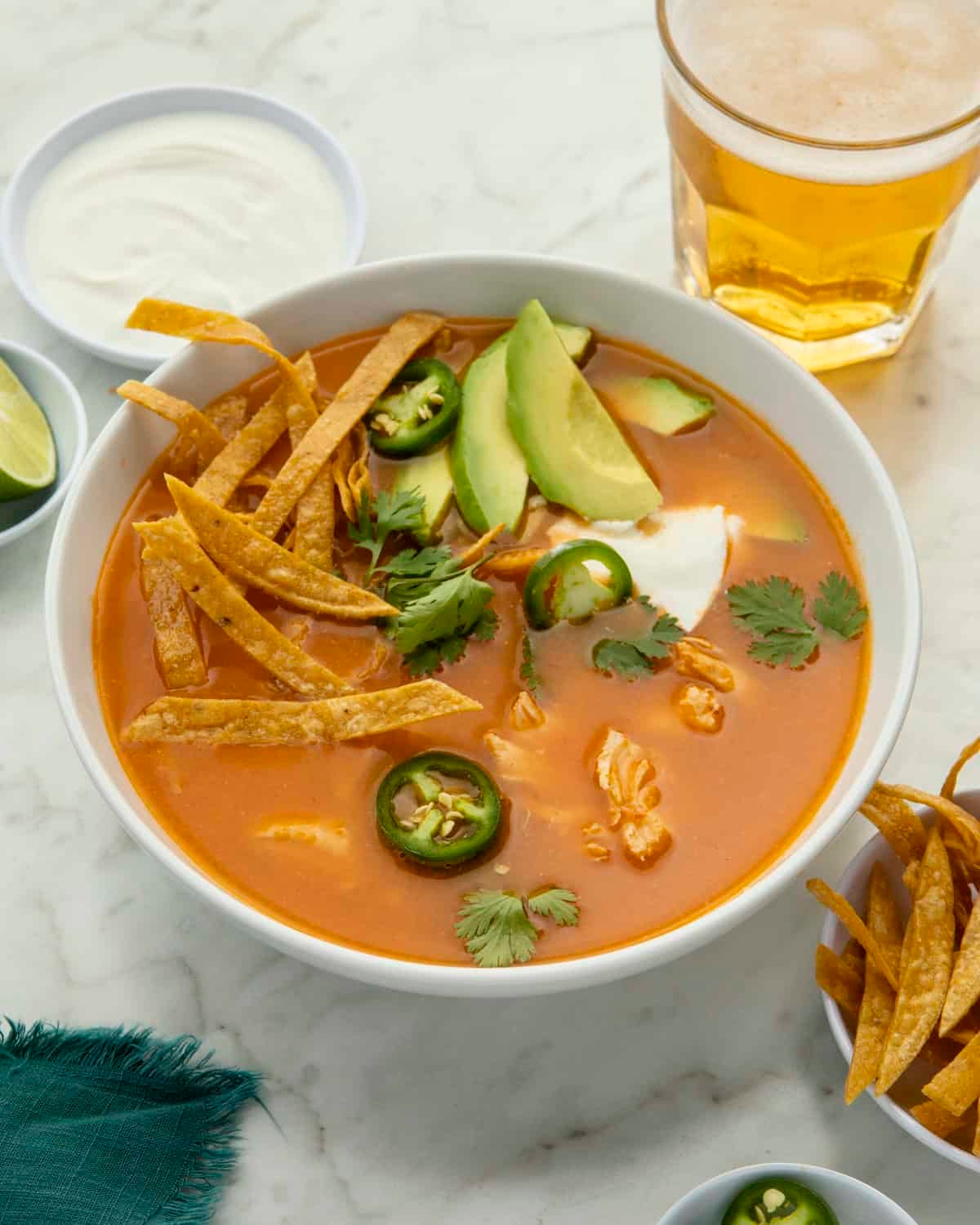 A bowl of tortilla soup with avocado, jalapeños, tortilla strips, and cilantro, served with a side of lime and a glass of beer