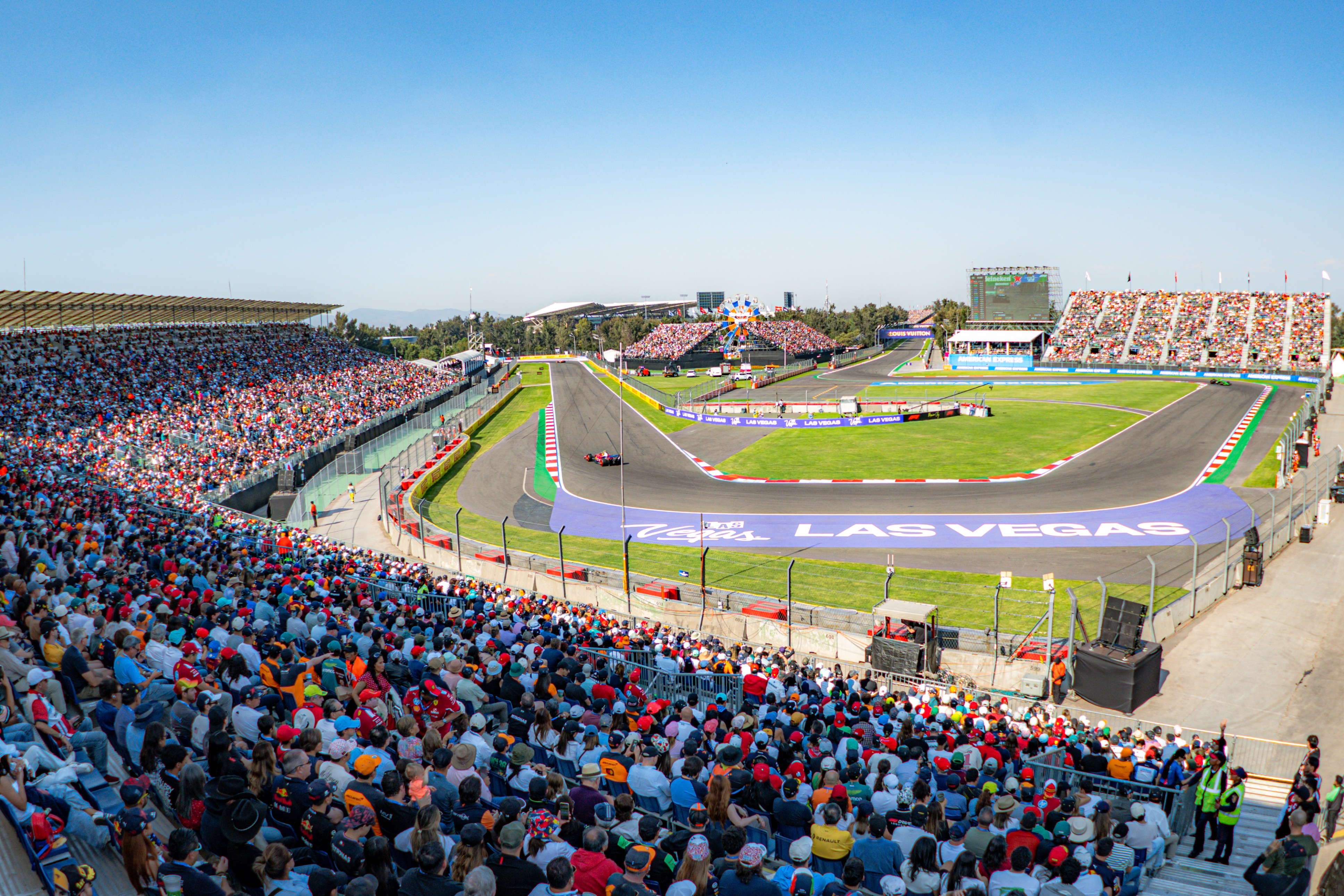 A packed stadium watches a car race on a bright day, featuring a turn with "Las Vegas" prominently displayed on the track