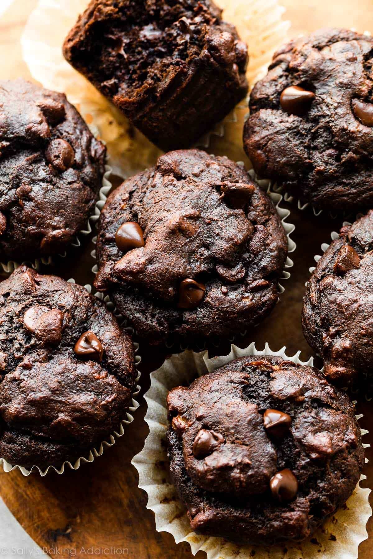 Chocolate muffins with chocolate chips are arranged on a wooden surface, one with a bite taken