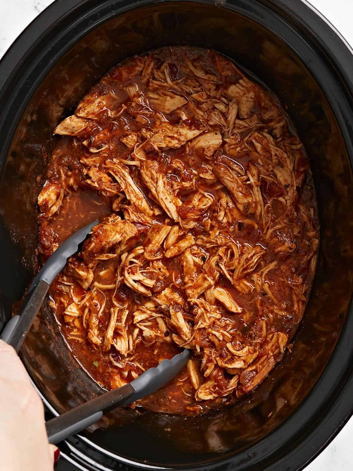 Shredded chicken in sauce inside a slow cooker, being served with tongs