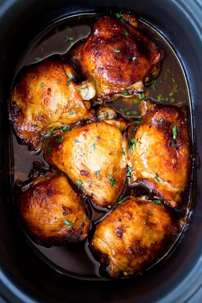 Cooked chicken thighs in a slow cooker, topped with herbs and surrounded by sauce