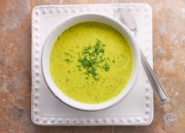 A bowl of creamy soup garnished with herbs, served on a square plate with a spoon on the side