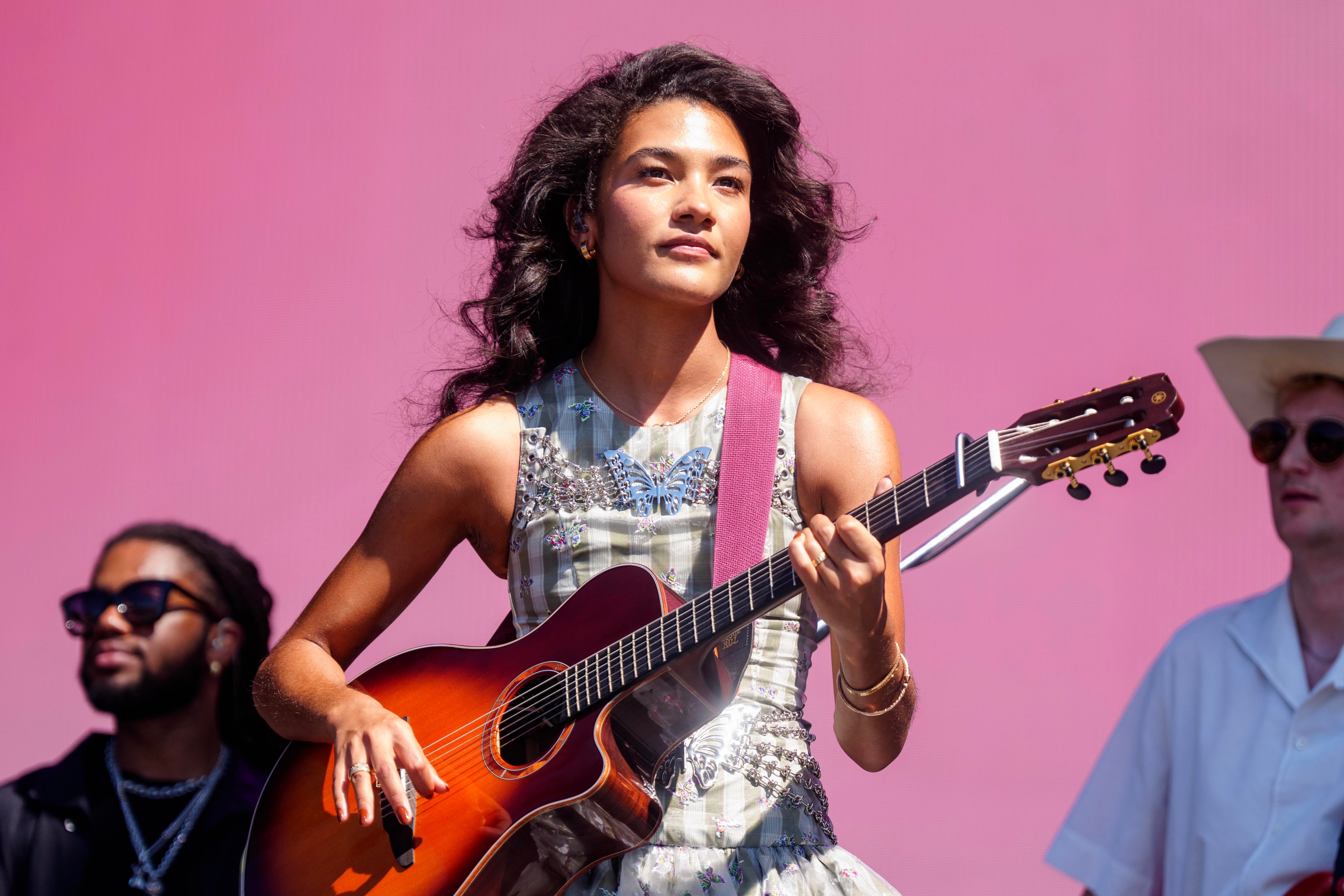 A person in a bejeweled dress plays an acoustic guitar on stage, with two others in the background, one wearing sunglasses and the other a hat