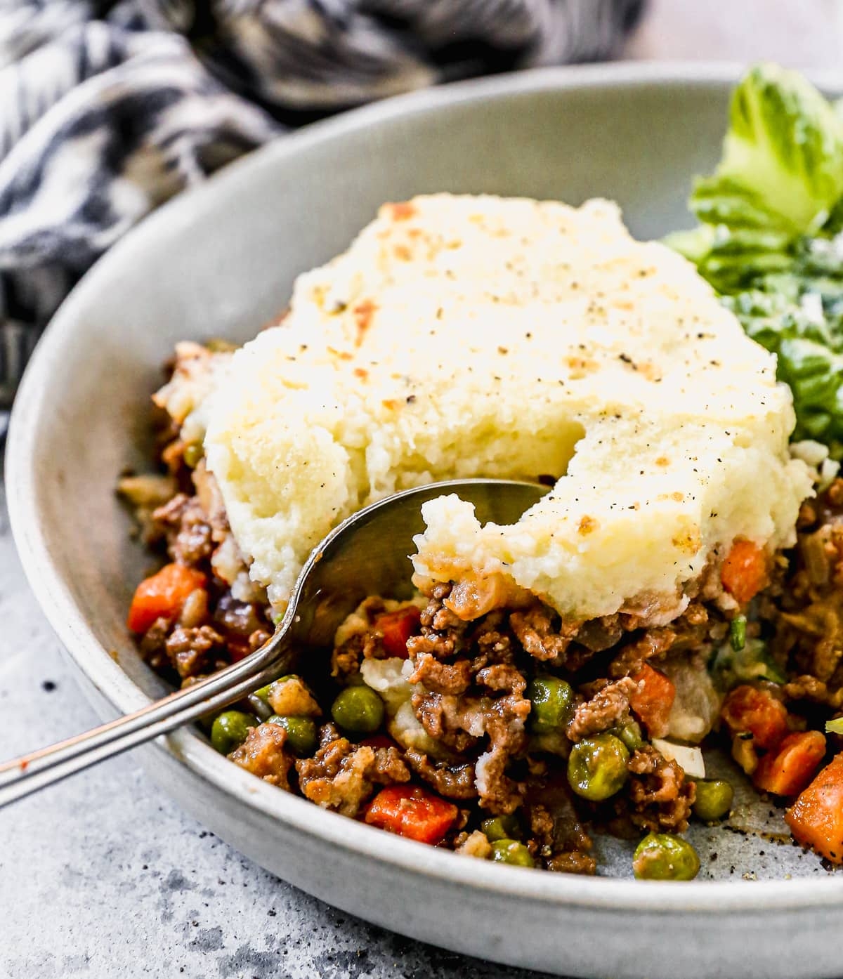 Shepherd's pie in a bowl with a spoon scooping mashed potatoes over a filling of minced meat, peas, and carrots