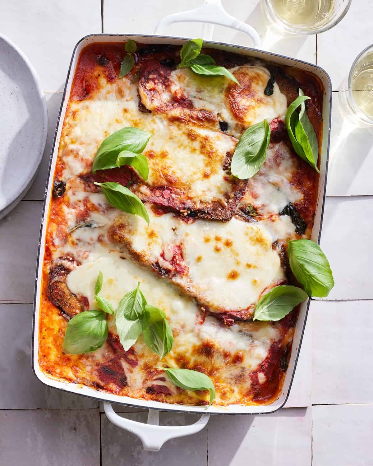Baked eggplant parmesan topped with melted cheese and fresh basil leaves in a rectangular dish. Plates and drinks nearby