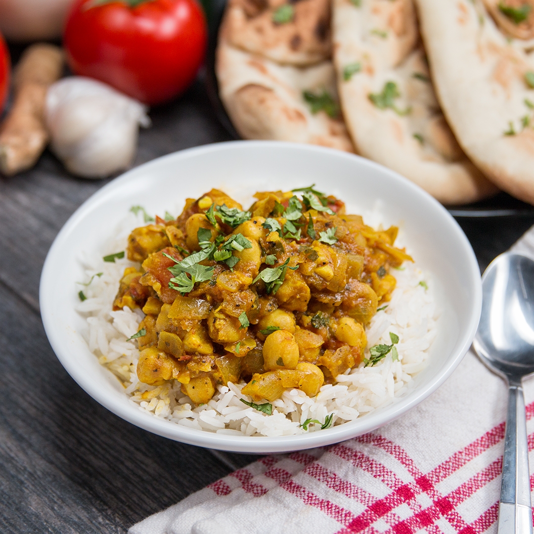 A bowl of chickpea curry with rice, garnished with herbs. Naan bread, garlic, and tomato are in the background on a wooden table