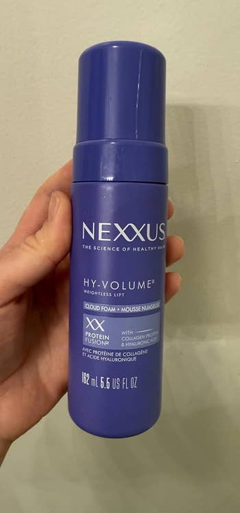 Hand holding a vessel of Nexxus Hy-Volume mousse, labeled with keratin macromolecule and collagen for weightless lift, 6.5 fl oz size
