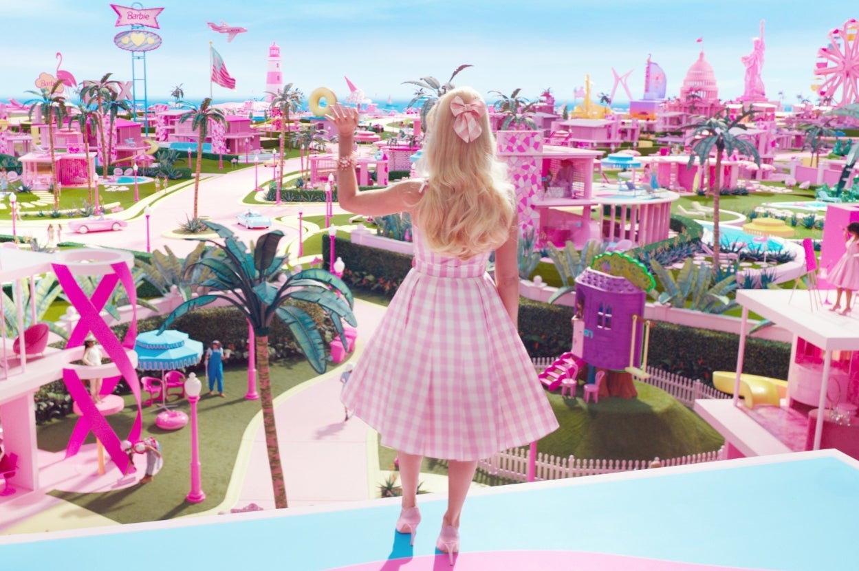 Margot Robbie as "Barbie" stands overlooking a vibrant town with whimsical architecture and playful design elements, hand raised