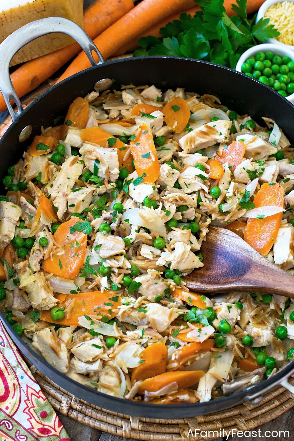 A skillet filled with a dish of shredded chicken, carrots, peas, and herbs, with a wooden spoon in view
