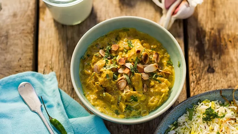 Bowl of curry topped with sliced almonds and herbs, next to a plate of rice on a wooden table
