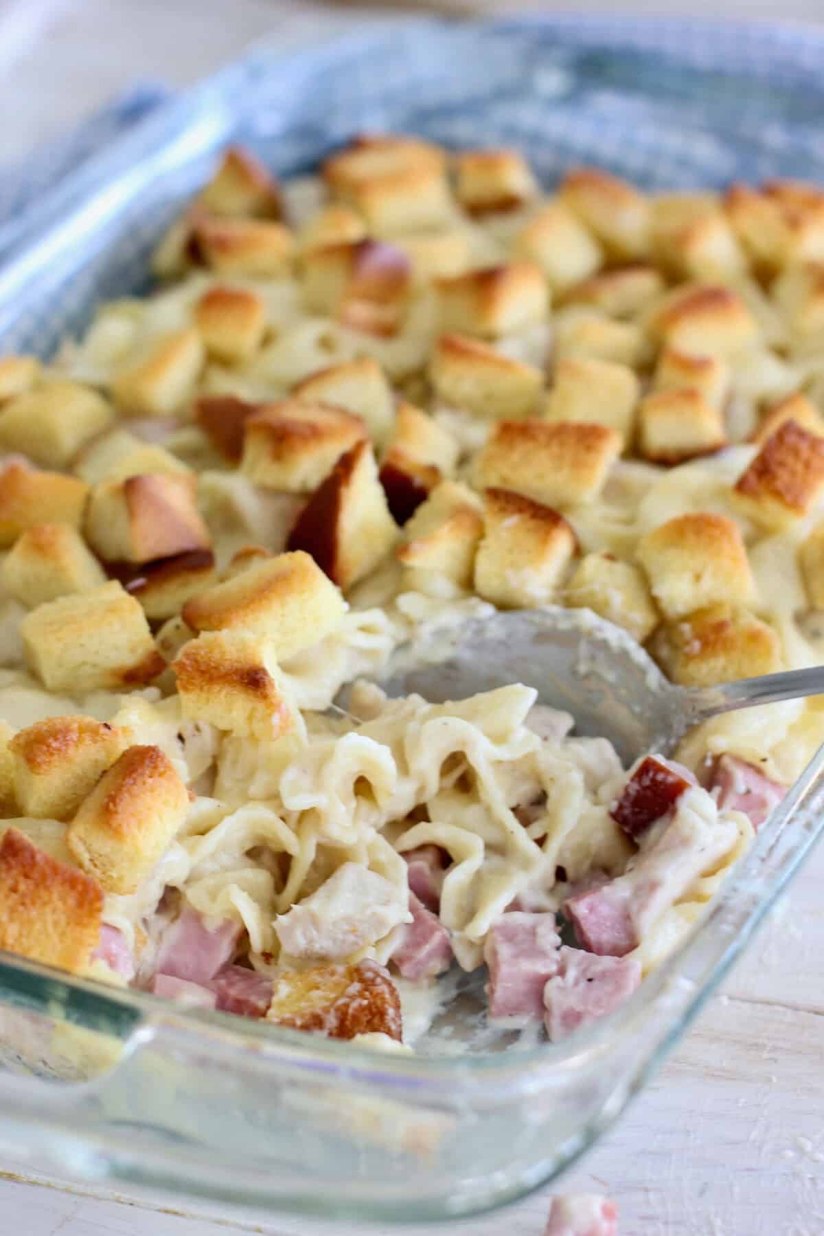 Baked pasta casserole with ham, creamy sauce, and crisp bread cubes in a glass dish. Spoon in the corner for serving