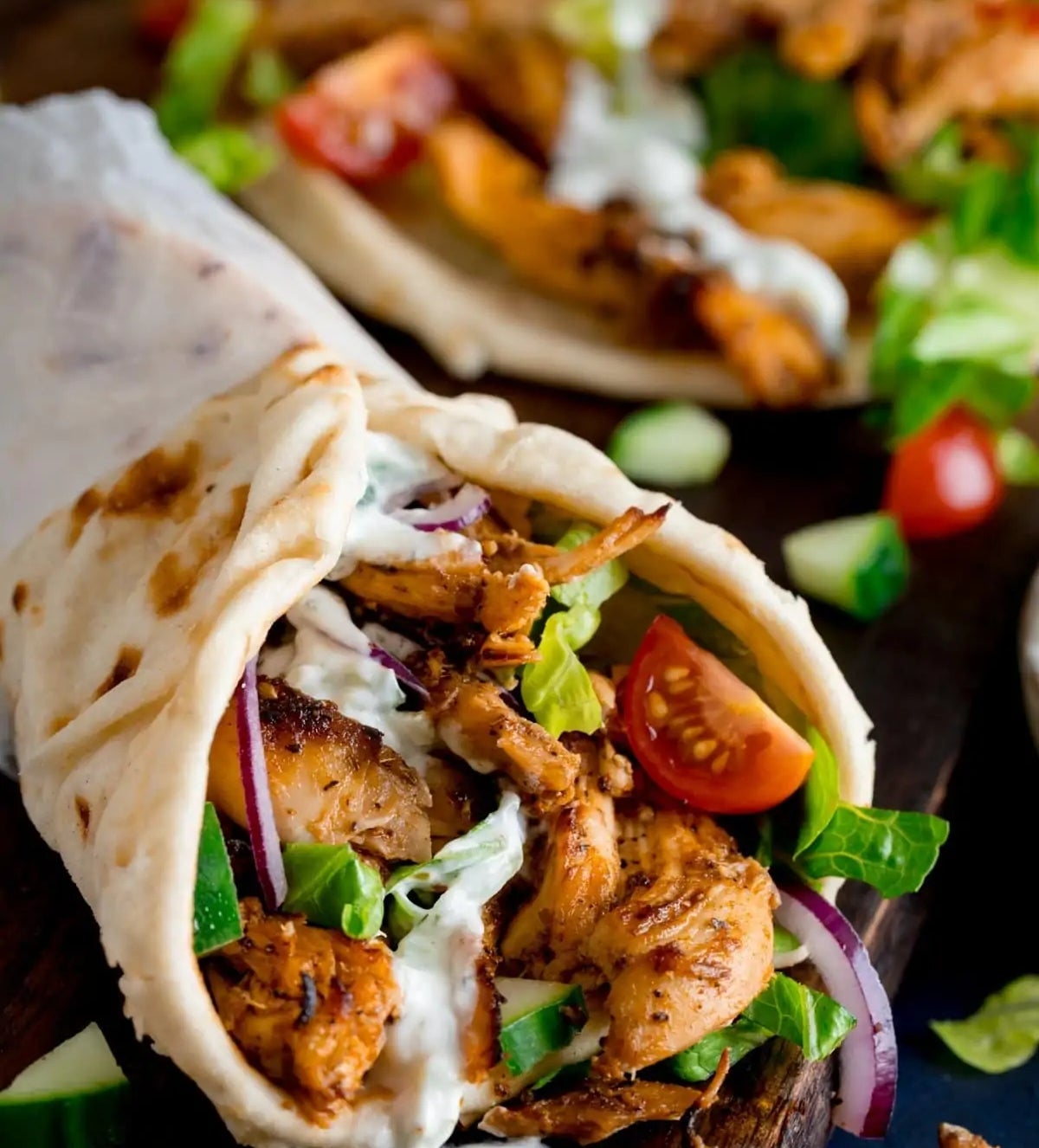 Close-up of a chicken wrap with grilled meat, fresh vegetables, and creamy sauce in a soft tortilla on a dark wooden surface