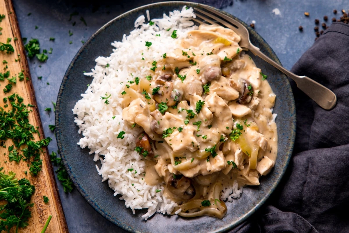 A plate of creamy chicken stroganoff with mushrooms and herbs served alongside white rice on a dark plate, garnished with fresh parsley