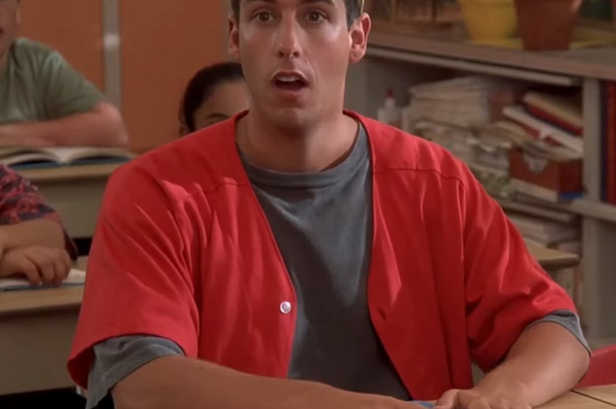 Person in a classroom, wearing a casual cap and open jacket over a t-shirt, sitting at a desk with an open book, looking surprised