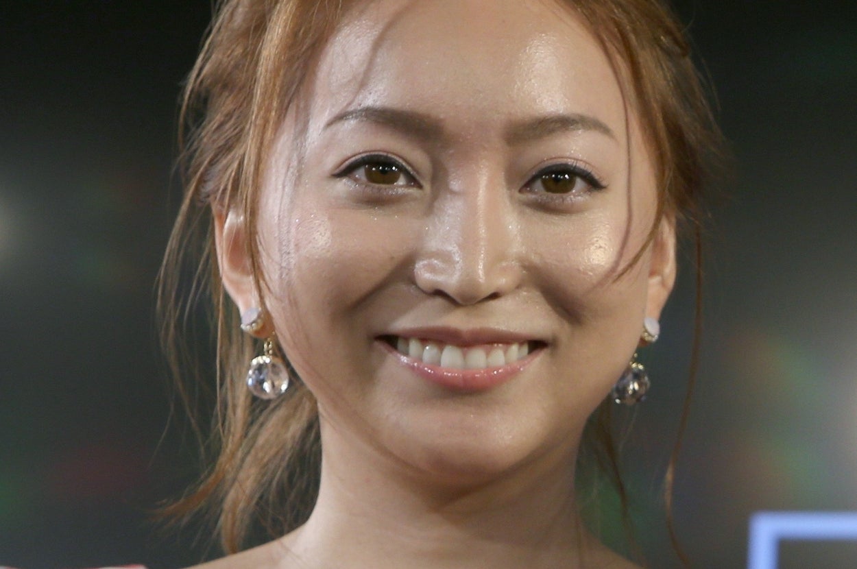 女性が笑顔でカメラに向かって立っている。アップスタイルの髪型とシンプルなイヤリングを身につけている。