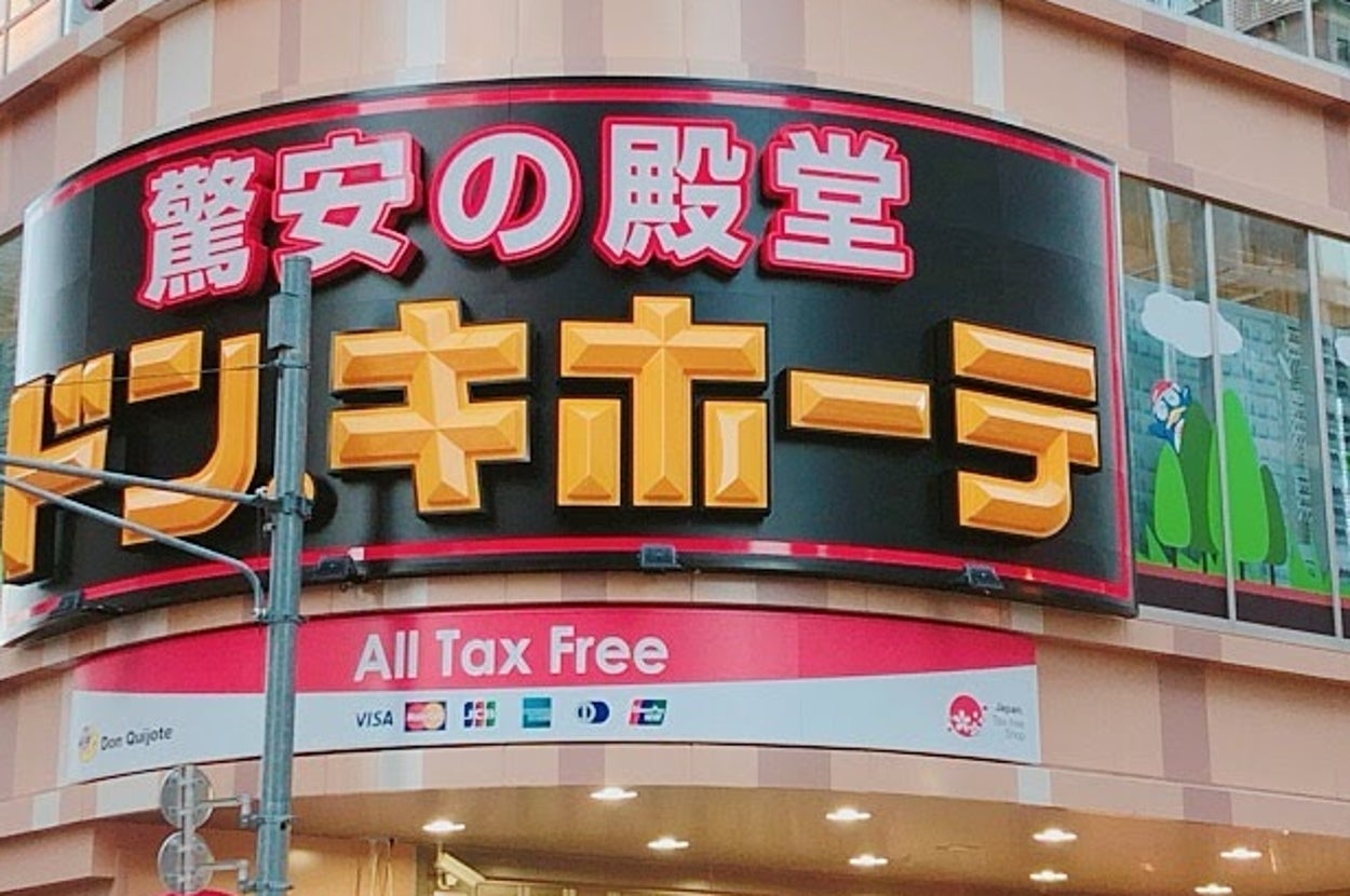 店の外観と通行人。目立つ看板が上に飾られ、賑やかな交差点に位置する店です。